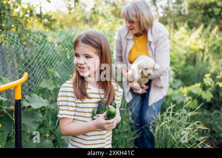 Das kleine Mädchen hält Gurken und lacht im Garten. Reife Frau hält goldene Retriever-Welpen draußen. Die süße Enkelin besucht ihre Großmutter. Stockfoto