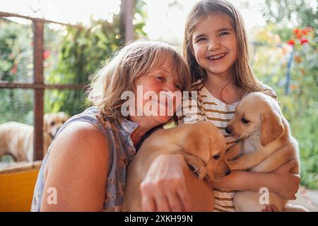 Reife Frau umarmt Welpen und ihre Enkelin. Das lächelnde kleine Mädchen hält einen goldenen Retriever. Niedliches Kind mit Großmutter kümmert sich um Haustiere. Rentner Stockfoto