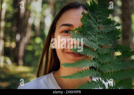 Nahporträt einer jungen Frau mit einem Farn in der Natur. Ein lächelndes Mädchen versteckt ihr Gesicht unter einem grünen Blatt im Wald. Brünette in einem weißen T-Shirt Stockfoto