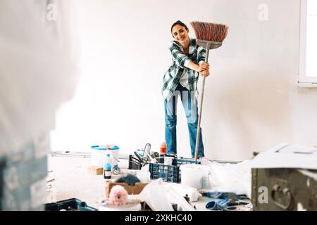 Eine junge Frau mit einer Bürste reinigt Schmutz im Zimmer. Mädchen in legeren Kleidern fegt den Boden mit Besen. Pause zwischen Reinigung. Baumaterialien Stockfoto