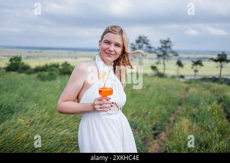 Eine junge Frau in einem weißen Kleid hält ein Glas mit einem orangen Cocktail auf einem Feld. Ein charmantes Mädchen genießt Entspannung und Natur. Eine lächelnde Blondine ist dri Stockfoto