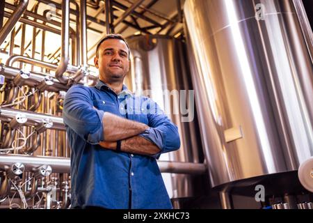 Ein junger, lächelnder Mann in lässiger Kleidung steht in der Werkstatt der Fabrik. Ein gutaussehender Arbeiter arbeitet in einer Fabrik. Ein Mann in einer Brauerei. Produktion e Stockfoto