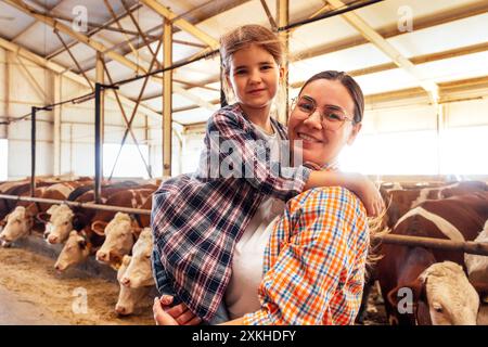 Eine junge Frau in lässiger Kleidung hält ihre kleine Tochter in den Armen in einem Kuhstall. Eine glückliche Bauernfamilie in einer Scheune. Ein süßes Mädchen und ihre Mutter sind s Stockfoto