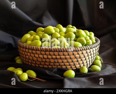 Runder Korb gefüllt mit grünen Oliven. Um den Korb herum gibt es auch Oliven. Hintergrund ist schwarzer Stoff. Stockfoto