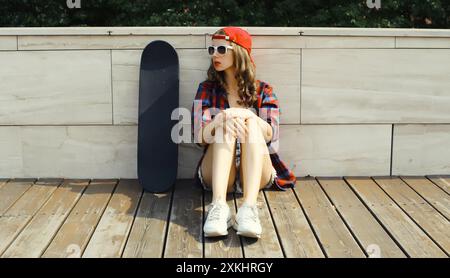 Stilvolle junge Frau, die mit Skateboard in legeren Kleidern sitzt und wegblickt, modernes Teenager-Mädchen, das in der Stadt posiert Stockfoto