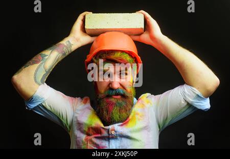 Maurer mit Ziegelstein. Maurer in orangefarbenem Schutzhelm und gefärbtem Hemd. Bärtiger Bauarbeiter im Schutzhelm mit Ziegel Stockfoto