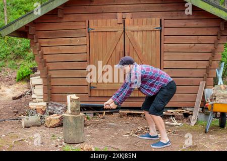 Mann im Hemd, der Holz mit Stahlaxt hackt, Landlandschaft mit Holzfäller, Brennholz für den Winter sammelt. Holzunterkunft und Wald im Hinterland Stockfoto