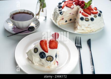 Pavlova-Kuchen oder Meringue-Dessert mit Beeren auf einem weißen Tisch und einer Tasse schwarzen Tee. Nahaufnahme, Fokus ausgewählt. Hochwertige Fotos Stockfoto