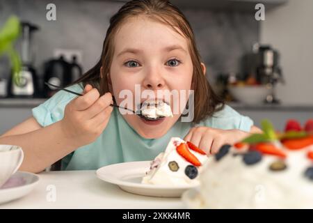 Das fröhliche kleine Mädchen isst Kuchen und steckt ihr ein großes Stück Dessert in den Mund. Ausgewählter Fokus. Hochwertige Fotos Stockfoto