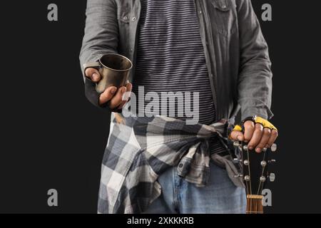 Reifer Obdachloser mit Tasse und Gitarre auf schwarzem Hintergrund, Nahaufnahme Stockfoto