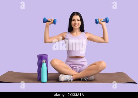 Sportliche junge Frau mit Kurzhanteln, die auf Matte vor Fliederhintergrund sitzen Stockfoto