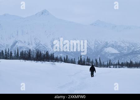 Mann, der im Schnee auf einem Weg zur Castner Cave mit Bäumen und Bergen im Hintergrund an einem kalten Wintertag in Alaska spaziert. Stockfoto