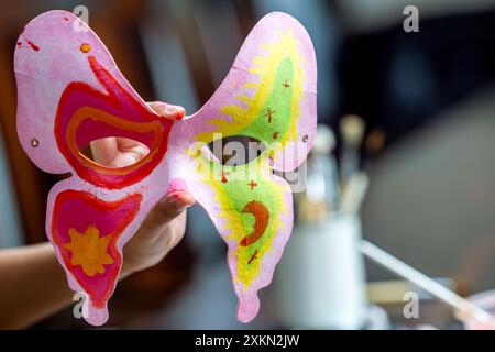 Eine Person hält eine Schmetterlingsmaske mit grünen und rosa Farben. Die Maske hat ein einzigartiges Design und ist mit vielen Details bemalt. Die Person scheint das zu tun Stockfoto