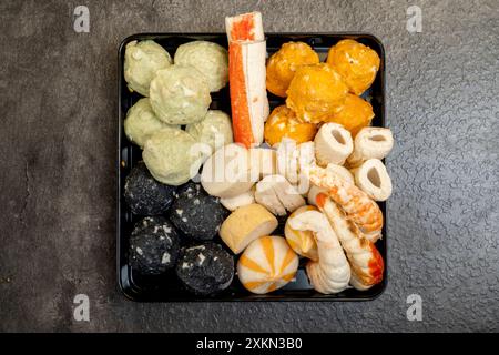 Ein schwarzes Tablett mit einer Vielzahl von Lebensmitteln, einschließlich einer Krabbe und einer grünen Kugel. Das Fach wird auf eine graue Oberfläche gestellt Stockfoto