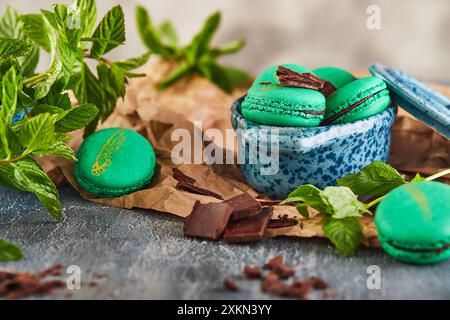 Grüne Macarons mit Schokoladenfüllung, umgeben von frischen Minzblättern und Schokoladenstückchen auf rustikalem Papier. Stockfoto