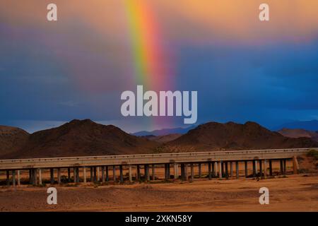 Leuchtender Regenbogen in der trockenen Wüste Kaliforniens mit leuchtenden Farben. Eine erhöhte Fahrbahn erstreckt sich über raues Gelände unter dramatischem Himmel Stockfoto