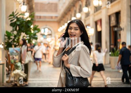 Eine attraktive, lächelnde asiatische Frau in modischer Kleidung spricht am Telefon, während sie in einer Einkaufsstraße in der Stadt läuft und den Camer ansieht Stockfoto