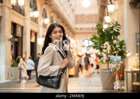 Eine attraktive, lächelnde asiatische Frau in modischer Kleidung spricht am Telefon, während sie in einer Einkaufsstraße in der Stadt läuft und den Camer ansieht Stockfoto