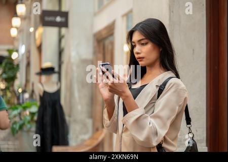 Eine attraktive asiatische Frau, die sich darauf konzentriert, Nachrichten auf ihrem Smartphone zu lesen, während sie vor einem Bekleidungsgeschäft in einer Einkaufsstraße in der Stadt steht. Stockfoto