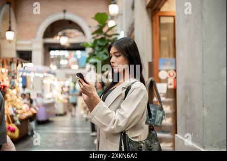 Eine attraktive asiatische Frau, die sich darauf konzentriert, Nachrichten auf ihrem Smartphone zu lesen, während sie vor einem Bekleidungsgeschäft in einer Einkaufsstraße in der Stadt steht. Stockfoto