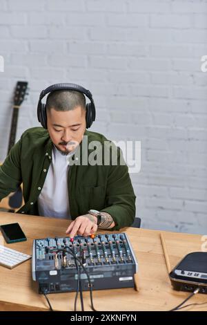 Ein hübscher asiatischer Mann mit Kopfhörern sitzt am Schreibtisch und mischt Musik mit einem Soundboard. Stockfoto