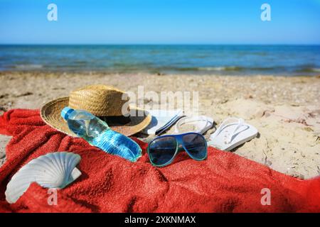 Badeurlaub am Strand mit rotem Handtuch, Wasserflasche, Strohhut, Sonnenbrille und Flipflops, Ostsee in Deutschland, Kopierraum, ausgewählter Fokus Stockfoto