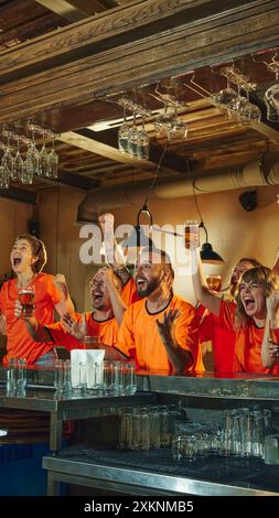 Freunde in orangefarbenen Hemden jubeln begeistert an der Bar, heben ihr Bier hoch und feiern den Sieg der Lieblingsteams. Stockfoto