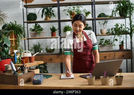Eine schöne asiatische Frau auf einer Schürze arbeitet in ihrem Werk und macht sich Notizen, während sie ihr kleines Unternehmen leitet. Stockfoto