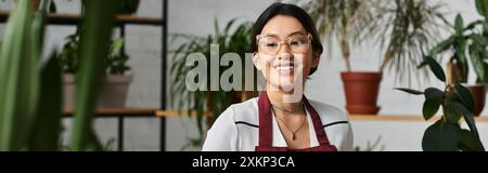 Eine lächelnde asiatische Frau mit Schürze und Brille steht in ihrem Pflanzenladen, umgeben von schönem Grün. Stockfoto