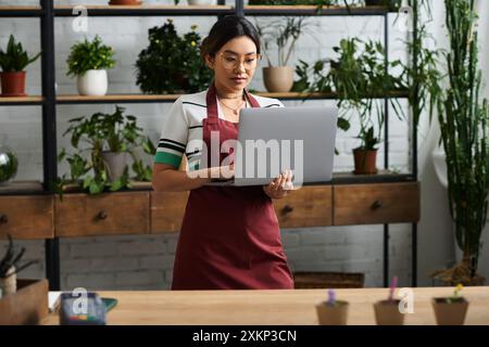 Eine junge asiatische Frau, die eine Schürze trägt, steht in ihrem Fabrikladen und nutzt ihren Laptop, um ihr Geschäft online zu verwalten. Stockfoto
