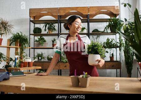 Eine schöne asiatische Frau in einer Schürze arbeitet an ihrem Ladentisch, hält eine Topfpflanze und steht bereit, Kunden zu bedienen. Stockfoto