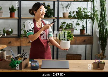 Eine Frau lächelt, während sie eine Topfpflanze in ihrem Blumenladen hält. Sie scheint am Telefon zu sprechen und nimmt wahrscheinlich eine Bestellung entgegen. Stockfoto
