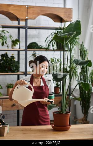 Eine asiatische Frau auf einer Schürze bewässert eine Pflanze in ihrem Pflanzenladen, umgeben von Grün und Regalen anderer Topfpflanzen. Stockfoto