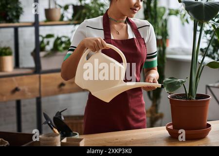 Eine beschnittene Frau mit Schürze bewässert eine Pflanze in ihrem Pflanzenladen. Stockfoto