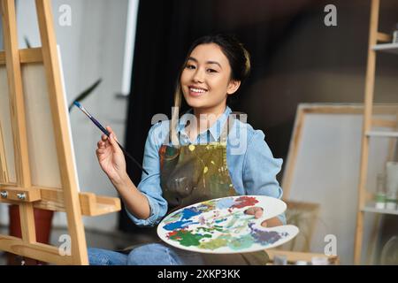 Eine glückliche asiatische Künstlerin malt in ihrer Werkstatt und hält einen Pinsel und eine Palette. Stockfoto