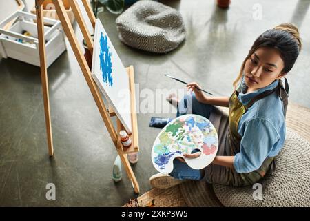 Eine junge asiatische Frau in einer Schürze sitzt auf dem Boden in einer Werkstatt und malt auf einer Leinwand. Stockfoto