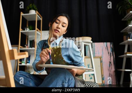 Eine asiatische Künstlerin, die eine Schürze trägt, sitzt in ihrem Atelier und mischt Farbe auf ihre Palette. Stockfoto