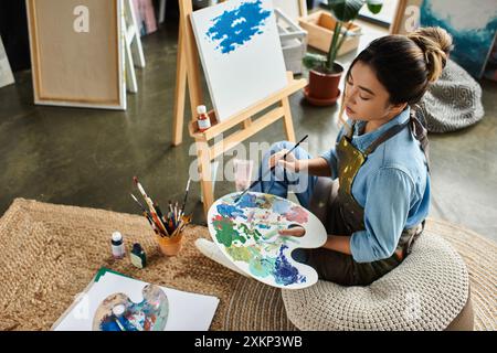 Eine junge asiatische Frau in einer Schürze sitzt auf einem Kissen in ihrer Werkstatt und mischt während sie malt intensiv Farben auf einer Palette. Stockfoto