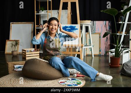 Eine junge asiatische Frau in einer Schürze sitzt auf einem Sitzsackstuhl in ihrem Kunststudio, lächelt und hält einen Pinsel. Stockfoto