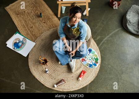 Eine junge asiatische Frau in einer Schürze sitzt auf einem runden Teppich, hält Pinsel und reflektiert ihr Kunstwerk. Stockfoto