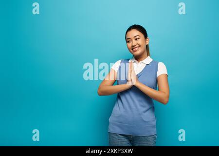 Eine junge asiatische Frau in eleganter Freizeitkleidung steht vor blauem Hintergrund, lächelnd und mit aneinander gefesselten Händen. Stockfoto