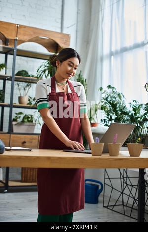 Eine schöne asiatische Frau mit Schürze arbeitet an einem Laptop in ihrem Werk, umgeben von Grün. Stockfoto