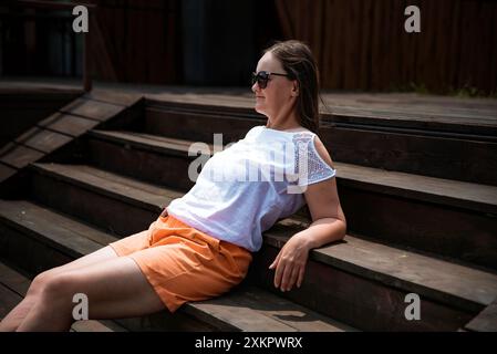 36 Jahre alte russin in orange Shorts und Sonnenbrille, die auf einem Balkon sitzt und am Sommertag genießt Stockfoto