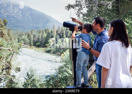 Vater und Sohn sehen die Berge und den Fluss durch ein Fernglas mit einem Münzempfänger an. Eine glückliche Familie von Touristen genießt die wunderschöne Landschaft. Stockfoto