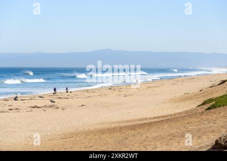 An einem nebeligen Herbsttag in Kalifornien fischen und spazieren die Leute an einem Sandstrand entlang. Große Wellen, die am Ufer brechen, sind in der Entfernung sichtbar. Stockfoto