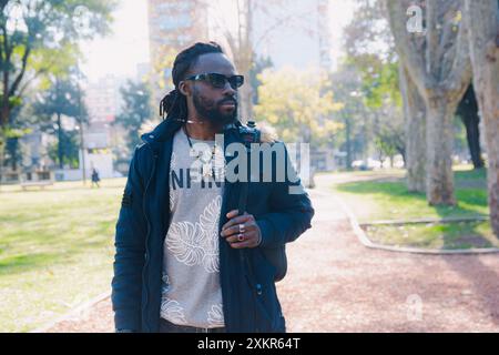 Ernstzunehmender junger schwarzer Mann, der im Park steht und zur Seite blickt, er trägt eine Sonnenbrille, hat Bart und Dreadlocks, er schaut sich mit nachdenklichem Gesicht um und Stockfoto