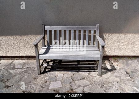 Eine hölzerne Gartenbank steht im Schatten an einer Hauswand. Der Hinterhof hat verrückte Pflaster. Stockfoto