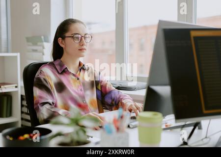 Junge kaukasische Programmiererin in gefleckter Bluse, die am Schreibtisch sitzt, während sie auf der Tastatur tippt und direkt auf den Computerbildschirm blickt Stockfoto