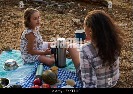 Mutter und Tochter teilen sich ein warmes Getränk, während sie draußen ein Picknick machen. Die Szene fängt einen Moment der Verbundenheit und Entspannung in einer natürlichen Umgebung ein. Stockfoto