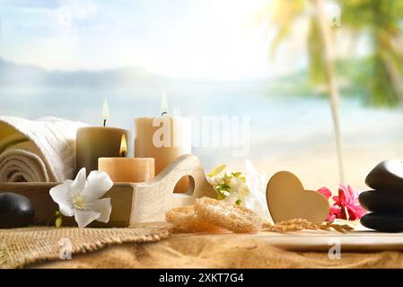 Wellness für Körper und Geist mit Produkten und dekorativen Elementen auf einem Holztisch mit Kerzen, Handtuch und Blumen mit Strand im Hintergrund. Vorn VI Stockfoto